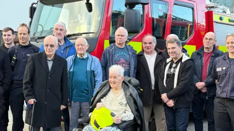 WPR David Hedger centre among old friends. David is in a wheelchair wearing a beige jumper with a black leather jacket over his shoulders and he is holding a yellow helmet.