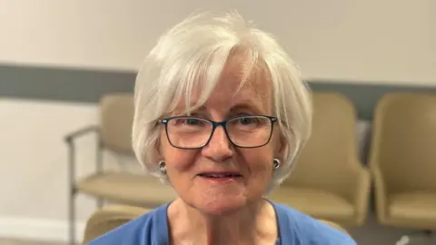 An older woman with short white hair and rectangular glasses. She is smiling and is wearing a blue top.
