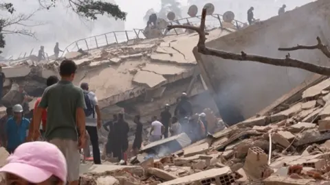 Survivors walk past a collapsed building with rubble strewn across the ground.