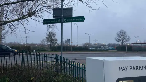 View of the Claret Car Park in Northampton. A pay machine is in the foreground and a sign shows directions to the nearby stadiums of Northampton Saints and Northampton Town