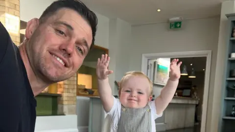 Glen, who has dark brown hair and wears a black T-shirt, leans forward to get into a selfie picture with Amber. She is sat in a high-chair and has her arms up in the air. She wears a white top and sage green dungarees, and the picture appears to have been taken in a restaurant. 