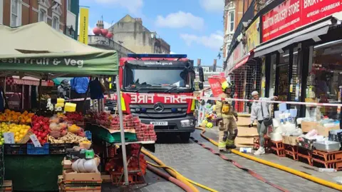 London Fire Brigade A fire engine in the street surrounded by stalls of fruit and vegetables.