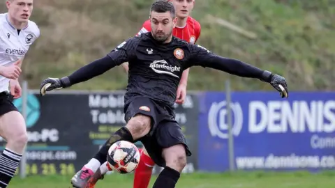 Pacemaker Aaron Hogg is kicking the ball in a football match. He is wearing an all black goalkeeper kit with gloves. Two other male players are in the background - one is running towards him from the left.