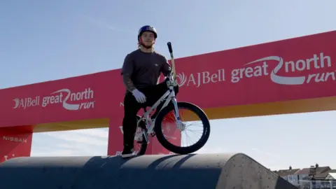 The BMX champion wearing a helmet sitting on a bike in front of the large, red Great North Run sign.