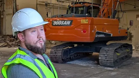 A man wearing a white hard hat and a yellow hi-vis vest standing in front of a large orange digger inside a warehouse-type building.