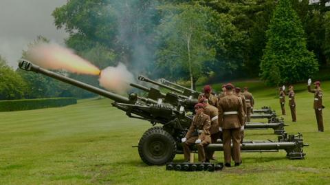 A row of green guns on wheels being fired by soldiers in ceremonial uniform