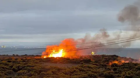 Fife Jammer Locations A fire at a colliery bing in Methil. The flames are orange with a plume of dark grey smoke coming off it. Green shrubbery is in the foreground.