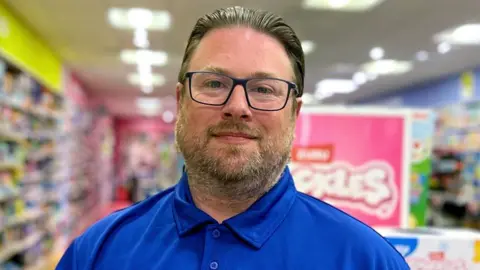 A head and shoulders shot of Lee James Adlam looking straight at the camera.
He wears the royal blue shirt which the uniform of The Entertainer and glasses. He has a beard and moustache. Behind him are the blurred outlines of boxes of toys. 