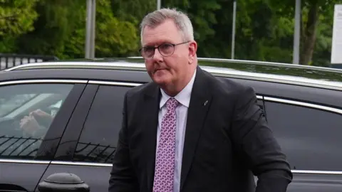 A man with mostly grey hair exiting a car. He is wearing a dark-coloured suit jacket, white shirt and pink tie. He is also wearing glasses. In the background, trees, lampposts and signs are visible
