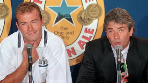 PA Media Alan Shearer sitting to the left of Kevin Keegan during a press conference. There are microphones in front of them both and Keegan is talking while Shearer looks off to the side and smiles. Shearer is wearing a white Newcastle United polo shirt while Keegan wears a black blazer and patterned tie. Shearer has dark blonde hair and Keegan has grey hair which is swept back. They are sitting in front of a large Newcastle Brown Ale advertisement.