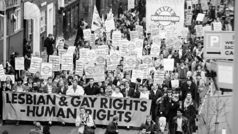 Getty Images A public demonstration showing crowds, holding placards and a large pro gay batter, marching down a road