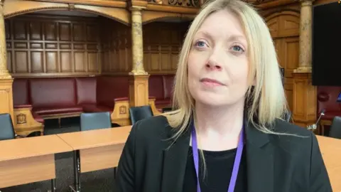 George Torr/BBC Head and shoulders shot of Natalie Hoy. She is wearing a black suit jacket and a purple lanyard around her neck.