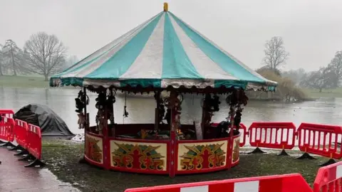 Peter Aitken A fairground stall can be seen next to Verulamium Lake in St Albans. It is surrounded by red barriers.