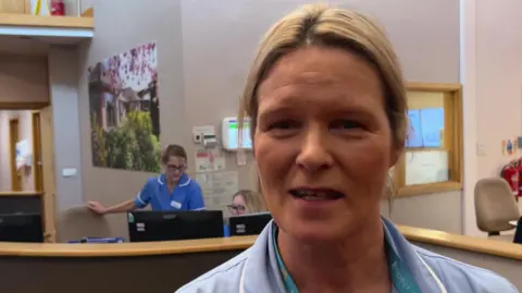 BBC/Nathan Turvey A picture of Nikki Smith wearing a blue uniform standing with hospice staff behind her at computer screens.