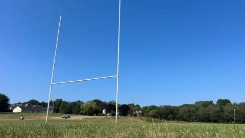 A low down image of rugby posts looking up at them on a clear blue day. The pitch is visible as well as trees in the background. 