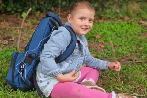 Jess John Lacie, 4, is sitting on the grass and smiling at the camera.
She is wearing pink leggings and a denim jacket and holding a stick in each hand. She has a large blue medical backpack on with a pipe going into a port. 