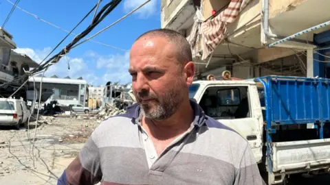 A man with very short hair and a greying beard stands on the street, looking off camera as he concentrates, his water truck behind him.