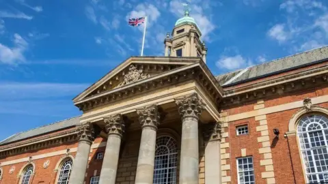 Brian Farmer/BBC A close up of Peterborough City Council town hall. The main building is brown brick, there is a 4 column roman style front and a flag pole with a union flag is above.