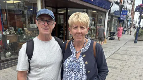 Jaimielee Rendall/BBC A man and woman are standing next to each other in Felixstowe town centre. They both have backpacks. The woman is wearing a blue jacket with old buttons and a blue and white shirt. The man is wearing a grey T-shirt, a blue hat and glasses. 