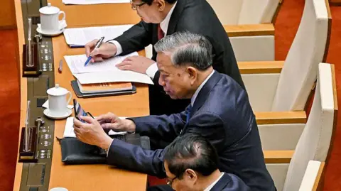 Getty Images Three men in suits sit at a long table, with documents and jugs of tea in front of them. The middle man is using a tablet.