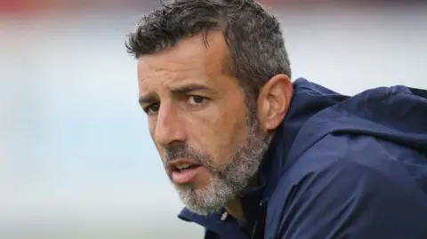 Getty Images Julio Arca during the pre-season friendly match between South Shields FC and Sunderland AFC at the First Cloud Arena on July 28, 2023 in Jarrow, England. He has grey hair and beard and brown eyes. He is wearing a blue coat.