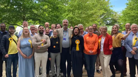 Emma Howgego/BBC A group of Liberal Democrat male and female councillors standing outside with trees behind them. They all appear to be shouting out and smiling. Third from the right at the front is the South Cambs Lib Dem MP Pippa Heylings.