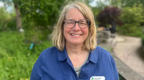 A woman smiling with blonde hair and glasses, wearing a blue shirt. She has a name badge on her left chest