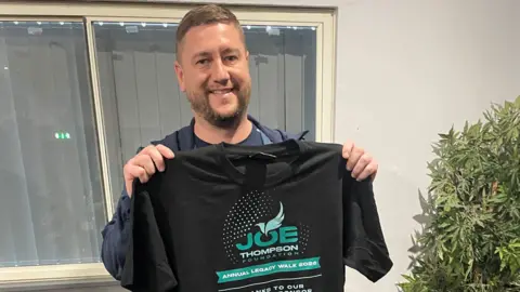A man standing inside by a plant holds up a black t-shirt which reads 'Joe Thompson Foundation Annual Legacy Walk 2026' in green lettering. He is smiling and has a short beard and cropped brown hair. 