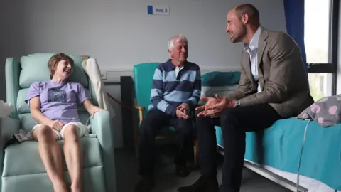 Reuters Prince William sat on a hospital bed while speaking to two patients who are sat on chairs. 