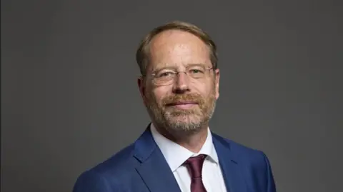 LDRS Andy MacNae, wearing glasses and a dark blue shirt and red tie, smiles for the camera in an official photo