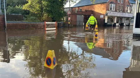 A flooded street with yellow cones in the water and a man in a high visibility jacket carrying two cones towards the opposite side of the street.