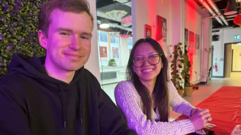 A man with brown hair smiling and a woman with dark hair and glasses smiling
