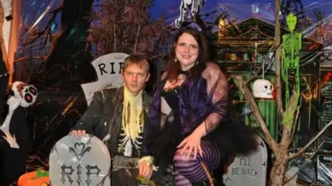 Gavin Hobbins, wearing a zombie costume, crouched in a set next to grave stones and skeletons, next to Kelly, wearing a purple witches costume