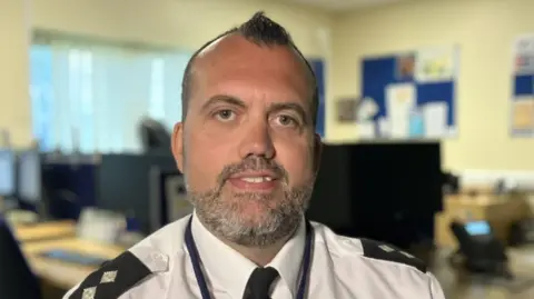 Tim Scott has short black hair and a beard. He is sitting in an office at a desk with computers behind him. He is wearing a white shirt with a black tie and epaulettes and is smiling.