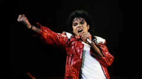 Getty Images Michael Jackson performs on stage, wearing a glittering red leather jacket over a white t-shirt. He holds a microphone to his mouth with one hand while the other is stretched out in front of him, fist clenched as he sings a note.