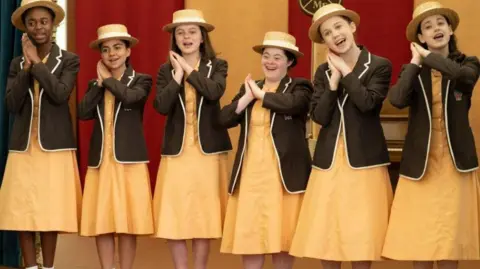 Six girls, including Ellie, in the boarding school uniform, line up in a row and appear to be singing and dancing. The girls are wearing yellow dresses and brown blazers. They have straw hats on and are in front of a red and yellow backdrop. 