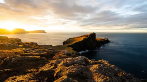 Chris Curry A dramatic coastal headland at sunset, with golden light hitting rocky cliffs that extend into the sea, and a lighthouse visible at the far end of the peninsula.