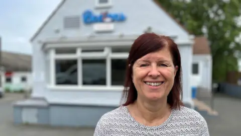 Kim Ashplant smiling at looking at the camera. She is wearing a light coloured top and has dark red hair. She is stood in front of the fish and chip shop which is blurred. 