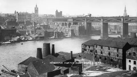 Newcastle Local Studies Library/Tyne Bridge A black and white view of a bridge across the Tyne and buildings either side of the river