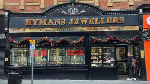 The outside of a jewellers shop, which has a black and gold frontage. It reads Hymans Jewellers above the door. In the windows are red and black displays of rings and necklaces.