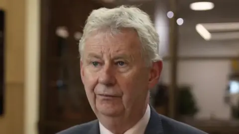 Edmund King is stood in an entrance foyer of the AA officers. He is wearing a dark suit, with a white shirt. He has short, grey hair.
