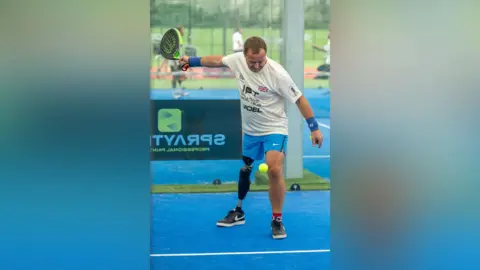 Andrew Simister Andrew Simister playing padel, on a bright blue padel court. An older man, Andrew is wearing a white t-shirt with black logos, blue shorts, and holds a padel raquet. He has a prosthetic leg.