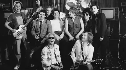 Getty Images A black-and-white photo of a group of nine people posing in front of a drum kit and amplifiers. Two in the front are seated, one holding an electric guitar, while the others stand behind them. 