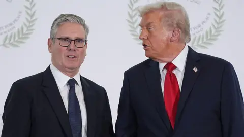 Sir Keir Starmer stands beside US President Donald Trump at an event, both wearing dark suits and ties, with Trump turned toward Starmer as if speaking while Starmer looks toward the camera.
