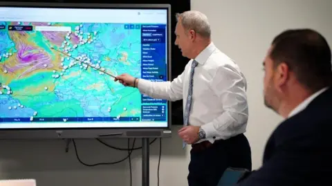 PA He uses a metal stick to point at a weather map on a screen. Another man in a black suit sits in the foreground watching. 