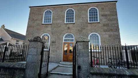 A picture of the stone building at dusk. There are arched windows with a set of wooden double doors and a path leading up to the entrance. A black metal wrought iron fence surrounds the perimeter.