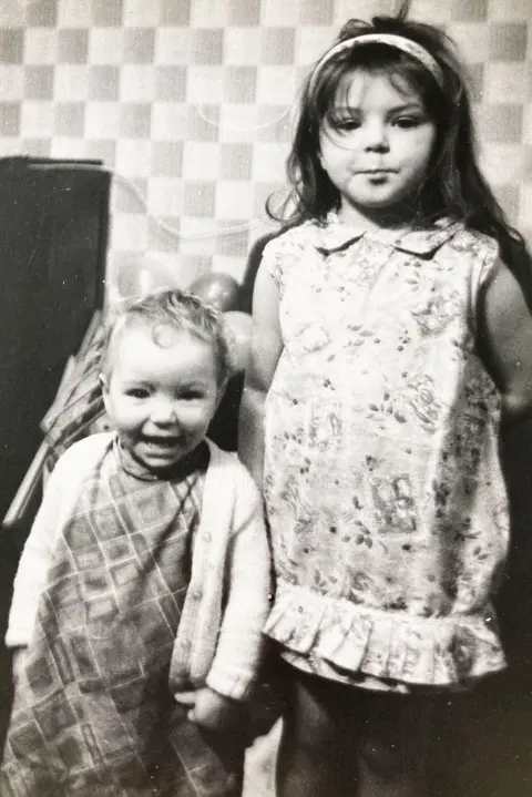 Theresa Fazzani/SWNS A black‑and‑white photograph of two young girls indoors. The older girl stands wearing a patterned dress with a headband, while a younger girl stands beside her grinning in a checked outfit and cardigan. Behind them is a wall with a distinctive square‑patterned wallpaper and some balloons.