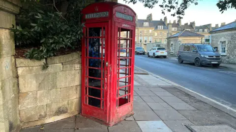 A red phonebox on the corner of a road. Cars are parked near by.