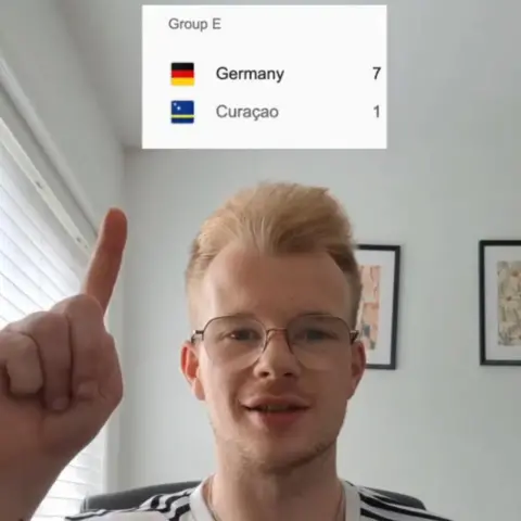 Tom Wilmott Tom is appearing in an Instagram post showing the Germany v Curacao result of 7-1 to the Germans in a box over his head. He has a German football top on.