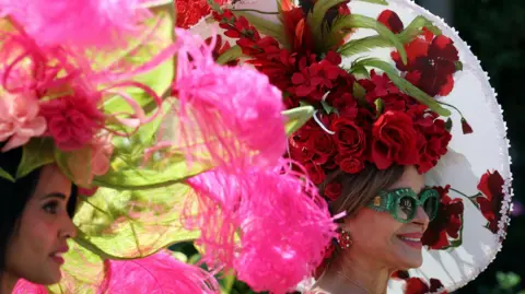 A woman with a hat which has large roses - either fake or real - attached to it, is pictured smiling for the camera, while another woman with other roses on her hat stands next to her.